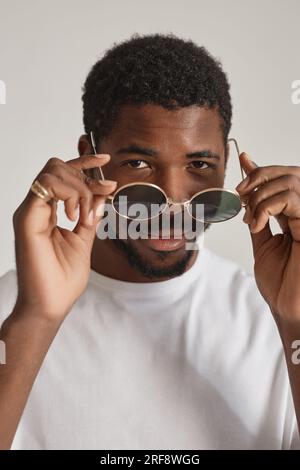 Ritratto verticale di un uomo nero sorridente che guarda gli occhiali da sole in uno studio bianco Foto Stock