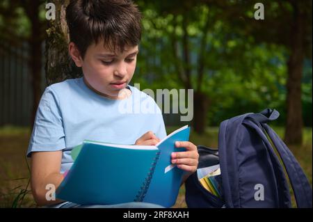 Primo piano di un bambino preadolescente ispanico dai capelli scuri, adorabile adolescente che scrive su un copybook, fa i compiti all'aperto. Ritorno a scuola conce Foto Stock