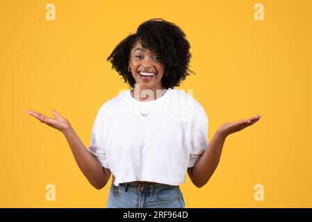 Happy sorprese la giovane donna nera riccia con una maglietta bianca, allarga le braccia ai lati, tieni lo spazio vuoto Foto Stock