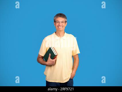 Uomo maturo felice che indossa occhiali che tiene libri su sfondo blu Foto Stock