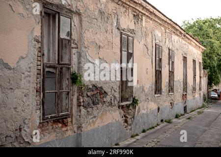 Esterno di un vecchio edificio abbandonato con muri in pietra sgretolanti e piccole finestre situate sulla strada di una piccola città Foto Stock
