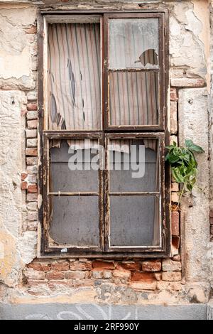 Esterno di un vecchio edificio abbandonato con muri in pietra sgretolanti e piccole finestre situate sulla strada di una piccola città Foto Stock