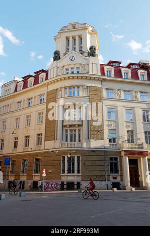 Via nel centro della città di Sofia, una signora di rosso fa una pedalata di fronte a un elegante e colorato edificio bancario, la Bulgaria. 1 agosto 2023. Foto Stock