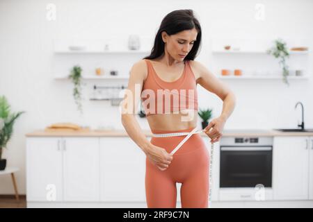 Donna attraente in buona salute misurazione della vita con nastro dopo l'allenamento a casa in abbigliamento sportivo yoga. Persona atletica che si dirige verso il miglioramento del benessere attraverso le abitudini alimentari e la forma fisica. Foto Stock