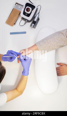 Il master unghie ritagliate rimuove la cuticola del cliente femminile con la chiavetta arancione, vista dall'alto. Lime, utensili per manicure sterilizzati in confezione artigianale su tavolo bianco Foto Stock