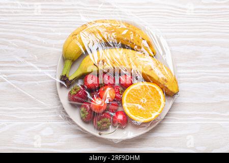 Frutta e frutti di bosco assortiti su piastra bianca avvolta in pellicola trasparente di plastica vista dall'alto su sfondo rustico bianco in legno Foto Stock