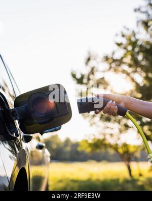 Giovane donna che collega il caricabatterie in un'auto elettrica nera, concetto di energia rinnovabile Foto Stock