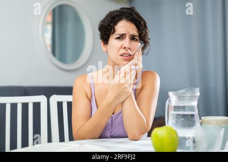 Giovane donna con mal di denti e guancia toccante, seduta a tavola Foto Stock