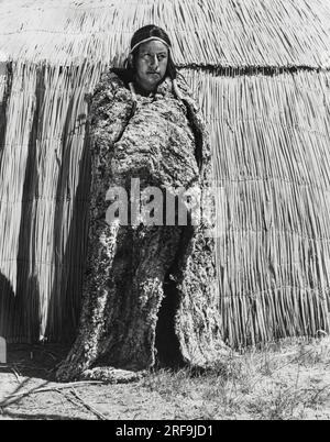 California: 1930 circa con il freddo, gli uomini dei nativi americani della California indossavano coperte di pelle per il calore. Questo Yokut indossa una coperta di pelle di coniglio fatta tessendo insieme strisce di pelle di coniglio. Per la maggior parte del resto dell'anno gli uomini indossavano solo un boccone o una pelle legata intorno alla vita. Foto Stock