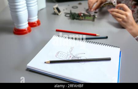 Robot disegna da bambino su un notebook con note sul pannello solare, mentre mani irriconoscibili di studenti che assemblano pezzi in una classe di robotica Foto Stock