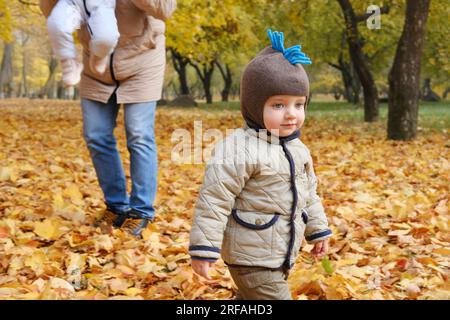 Il padre passa del tempo con i bambini nel parco autunnale. Il padre porta il bambino tra le braccia, il suo figlioletto cammina da solo. Foto orizzontale Foto Stock