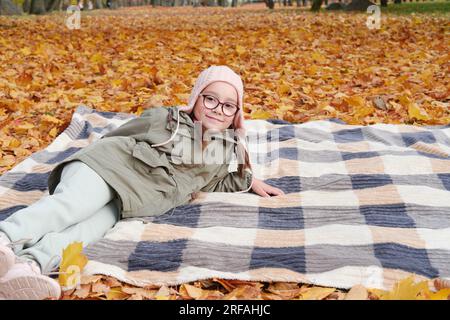 Bambina con una giacca autunnale, un cappello rosa e occhiali giace su una coperta in un parco autunnale. La ragazza sorride e guarda la telecamera. Foto orizzontale Foto Stock