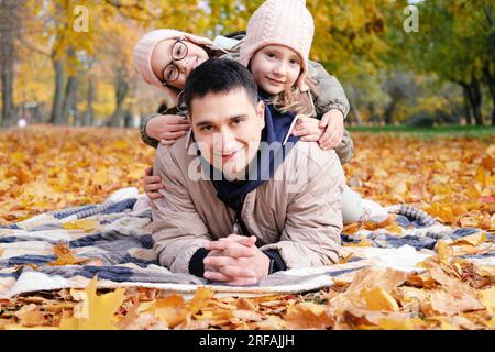 Giornata in famiglia nel parco autunnale. Le ragazze gemelle giacciono sulla schiena del padre. Padre e sorelle sorridono e guardano la telecamera. Foto orizzontale Foto Stock