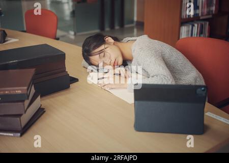 Studentessa stanca con gli occhiali sdraiati sui libri in biblioteca. Foto Stock