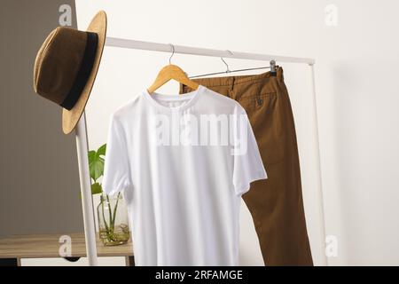 T-shirt bianca, cappello e pantaloni marroni appesi alla barra appendiabiti con spazio per le copie su sfondo bianco Foto Stock