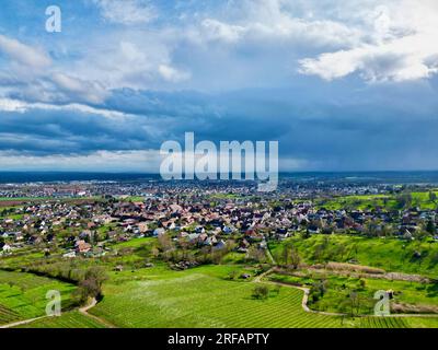 Uffholtz Winter Wonderland: I vigneti verdeggianti abbracciano un pittoresco villaggio tra cieli tempestosi, affacciati sulle pianure verdi alsaziane e sulle montagne Foto Stock