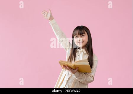 Una giovane ragazza asiatica allegra e luminosa tiene in mano un libro aperto e alza la mano mentre si trova su uno sfondo rosa isolato. askin Foto Stock