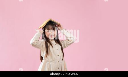 Una giovane ragazza asiatica sorridente e adorabile si mette in testa un libro mentre si trova su uno sfondo rosa isolato. imparare, ragazzo felice, educatore Foto Stock