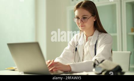 Medico femminile concentrato siede a tavolo in ospedale e lavora su portatile online Foto Stock