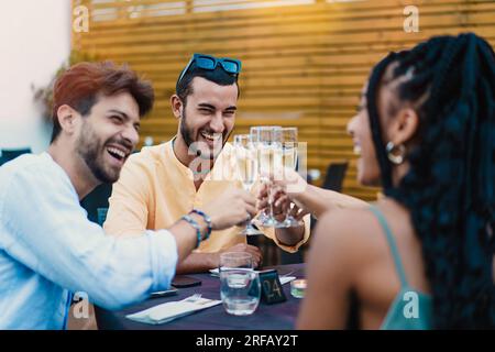 Un giovane alla moda con occhiali da sole e barba che conduce un brindisi con flauti di champagne tra i suoi diversi amici in un ristorante, incarnando gioia e camma Foto Stock