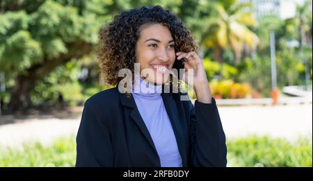 Ridendo donna d'affari latinoamericana che parla al telefono all'aperto in città d'estate Foto Stock