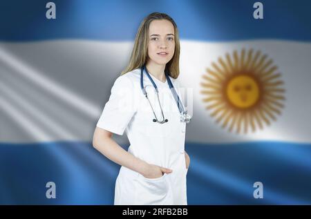 Una giovane medico donna con un mantello bianco e uno stetoscopio si trova sullo sfondo della bandiera argentina. Foto Stock