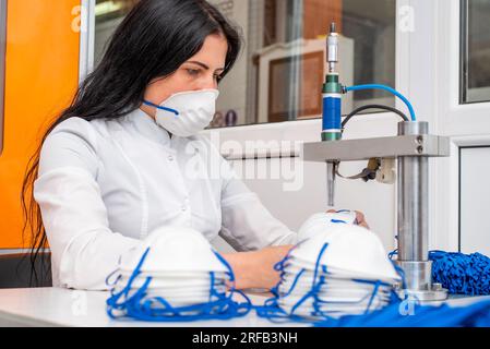 La ragazza lavora alla macchina per la fabbricazione di maschere mediche con nanofibra e saldare i loop a loro con ultrasuoni. Coronovirus e Covid- Foto Stock