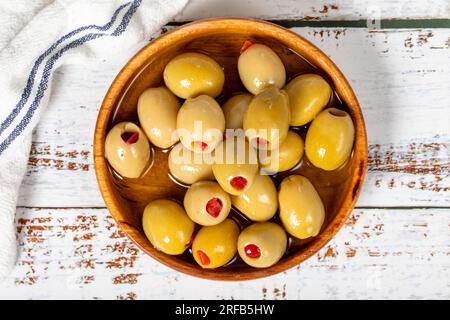 Olive verdi ripiene di pepe in un recipiente di legno. Olive su fondo di legno bianco. Cucina mediterranea. cibo vegano. Vista dall'alto Foto Stock