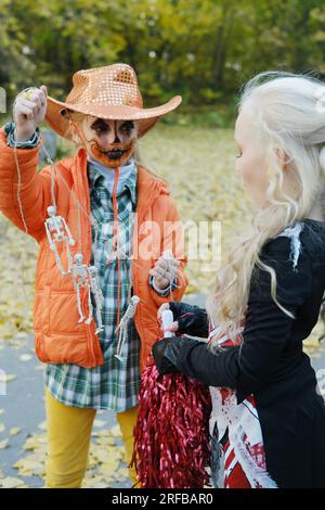 Bambina con una giacca arancione e un cappello da cowboy mostra alla sua amica in costume da cheerleader i suoi scheletri su una corda. Festa di Halloween. Pho verticale Foto Stock