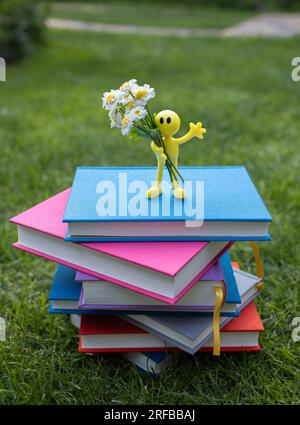 una pila di libri multicolore giace sull'erba, su di essa si erge una statuetta gialla umana ricordo con fiori di camomilla. biglietto per l'insegnante. concetto di educati Foto Stock