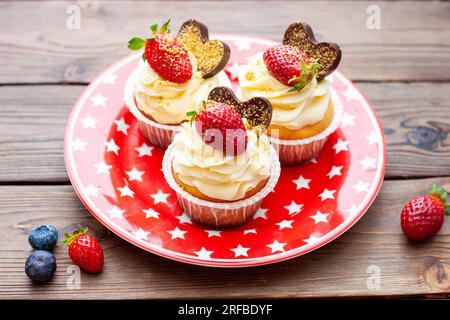 Set di cupcake alla vaniglia decorati con fragole fresche, mirtilli, cuori di cioccolato e spruzzi dorati su piatto rosso stellato Foto Stock