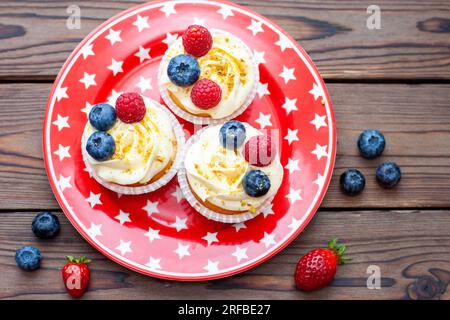 Set di cupcake alla vaniglia decorati con fragole fresche, mirtilli, cuori di cioccolato e spruzzi dorati su piatto rosso stellato Foto Stock
