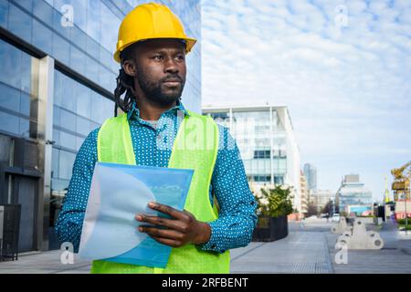 ritratto di uomo nero con barba e dreadlock, architetto con elmetto e gilet di sicurezza gialli, in piedi all'aperto pensando di tenere una cartella, spazio per copiare. Foto Stock