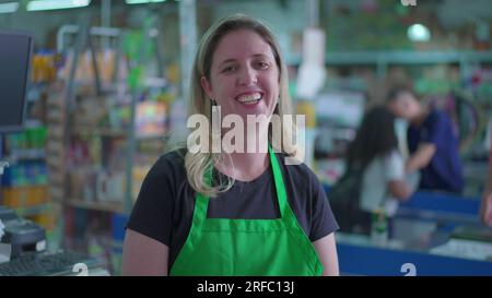 Un gioioso personale femminile del supermercato sorridente davanti alla macchina fotografica che indossa grembiule verde con gli acquirenti sullo sfondo al momento del check-out della cassa Foto Stock