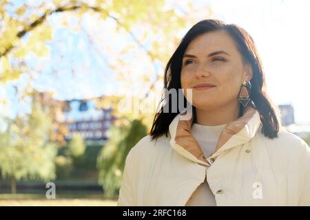 Bella donna bruna con lentiggini sul viso sorride e guarda in lontananza. Giovane donna che posa nel parco autunnale con il tempo soleggiato. Orizzontale Foto Stock