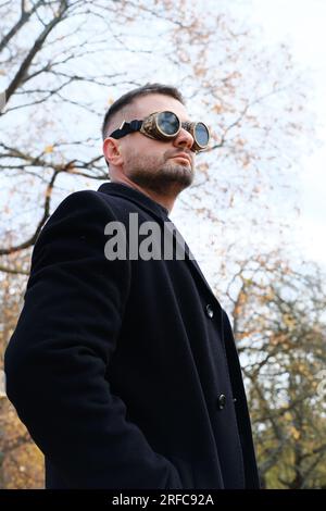 Un uomo con abito da lavoro, cappotto e occhiali steampunk si pone in un parco autunnale. Foto verticale Foto Stock