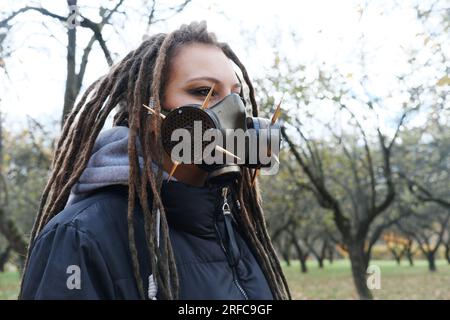 Ritratto di una donna in una giacca nera con dreadlock e una maschera antigas con spike. Donna che posa in un parco autunnale. Foto orizzontale Foto Stock