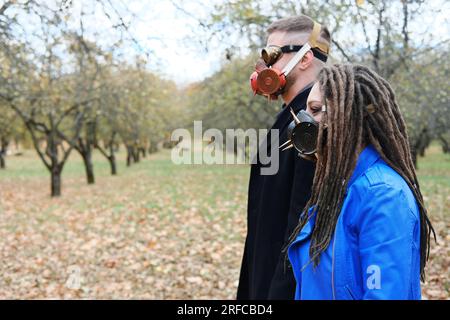 Una donna con dreadlock e una maschera a gas e un uomo con occhiali steampunk e una maschera a gas posa in un parco autunnale. Concetto di disastro ecologico. Orizzontale Foto Stock