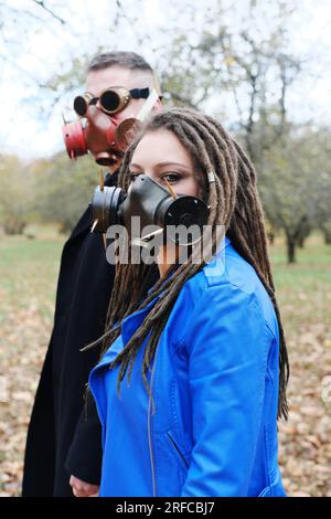 Una donna con dreadlock e una maschera a gas e un uomo con occhiali steampunk e una maschera a gas posa in un parco autunnale. Concetto di disastro ecologico. Verticale p Foto Stock