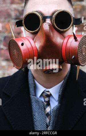 Un uomo con un cappotto nero, una tuta da lavoro, una maschera antigas e occhiali steampunk si pone contro un muro di mattoni. Foto verticale Foto Stock