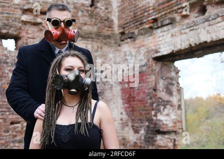 Due persone in abiti neri e maschere antigas si posano sullo sfondo di un muro di mattoni distrutto. Occhiali steampunk su un uomo. Foto orizzontale Foto Stock