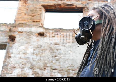 Sopravvivenza femminile post-apocalisse. Giovane donna sopravvissuta pandemica in maschera a gas che guarda di fronte a lei sullo sfondo con area di guerra apocalittica arou Foto Stock