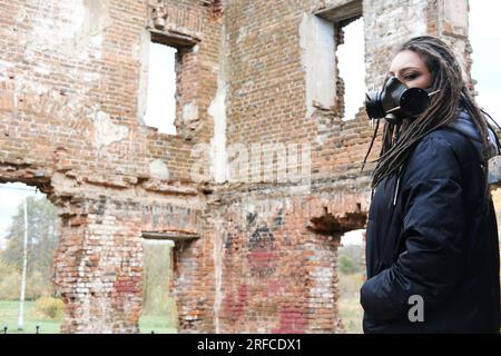 Sopravvivenza femminile post-apocalisse. Giovane donna sopravvissuta pandemica in maschera a gas che guarda di fronte a lei sullo sfondo con area di guerra apocalittica arou Foto Stock