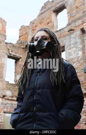 Sopravvivenza femminile post-apocalisse. Giovane donna sopravvissuta pandemica in maschera a gas che guarda di fronte a lei sullo sfondo con area di guerra apocalittica arou Foto Stock