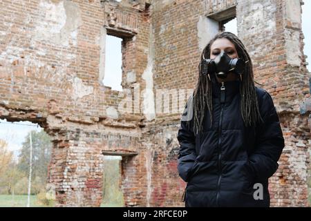 Sopravvivenza femminile post-apocalisse. Giovane donna sopravvissuta pandemica in maschera a gas che guarda di fronte a lei sullo sfondo con area di guerra apocalittica arou Foto Stock