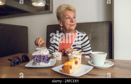 Donna anziana attraente e gioiosa che beve caffè mentre si siede in un bar. Ritratto di donna matura in una caffetteria. Torte colorate e caffè su un tavolo. Foto Stock