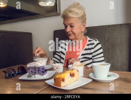 Donna anziana attraente e gioiosa che beve caffè mentre si siede in un bar. Ritratto di donna matura in una caffetteria. Torte colorate e caffè su un tavolo. Foto Stock