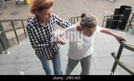 La donna anziana e la figlia di mezza età camminano nel cortile di un alto edificio. L'infermiera aiuta un vecchio pensionato a salire sulle scale all'esterno. Operatore sanitario Foto Stock