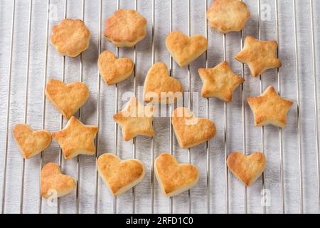 Biscotti al formaggio cottage appena sfornati a forma di stelle e cuori su una griglia d'acciaio, vista dall'alto. Fase di cottura di deliziosi biscotti fatti in casa. Foto Stock