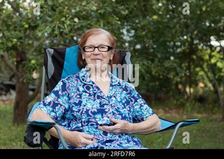 Ritratto della nonna anziana sorridente in giardino a casa. l'anziana si sta riposando seduto sulla sedia, guardando la macchina fotografica, il frutteto sullo sfondo. Somma Foto Stock
